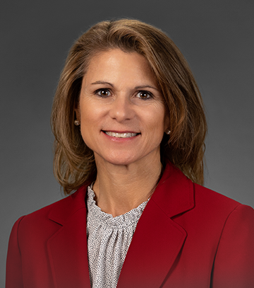 A professional headshot of Brenda Mackey, a smiling woman with shoulder length brown hear wearing a blouse and a red blazer, sitting against a light gray background. The subject is facing the camera with a friendly and approachable expression.