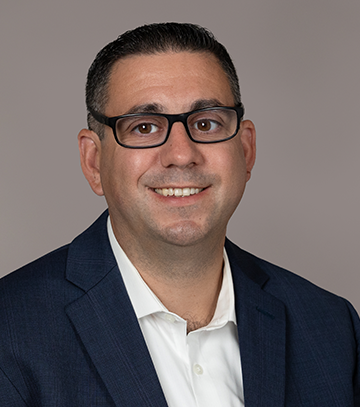 A professional headshot of David Semenza, a smiling man wearing a white collared shirt with a navy blue blazer, sitting against a light gray background. The subject is facing the camera with a friendly and approachable expression.
