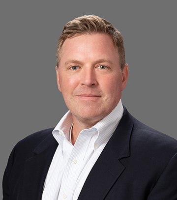 A professional headshot of Jeff Rathkamp, a smiling man wearing a white collared shirt and a black blazer, sitting against a light gray background. The subject is facing the camera with a friendly and approachable expression.